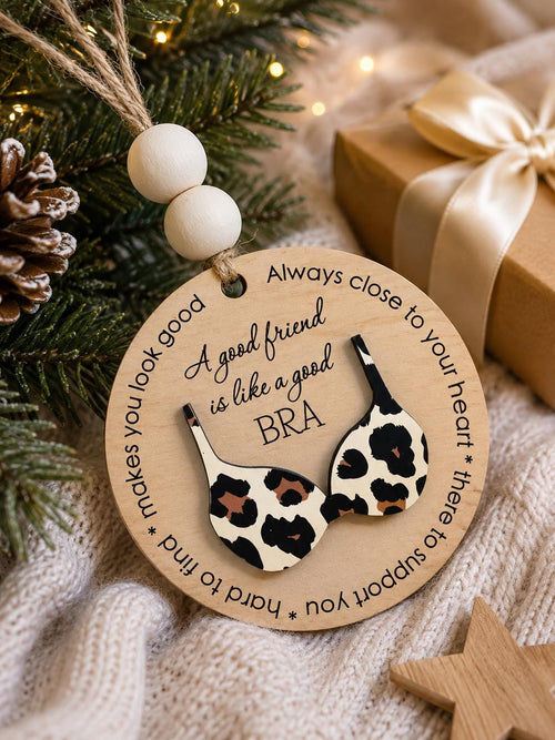 May include: A round wooden ornament featuring a leopard print bra cutout and the text: A good friend is like a good BRA, Always close to your heart, hard to find, makes you look good. It includes two white wooden beads and a jute twine hanger.